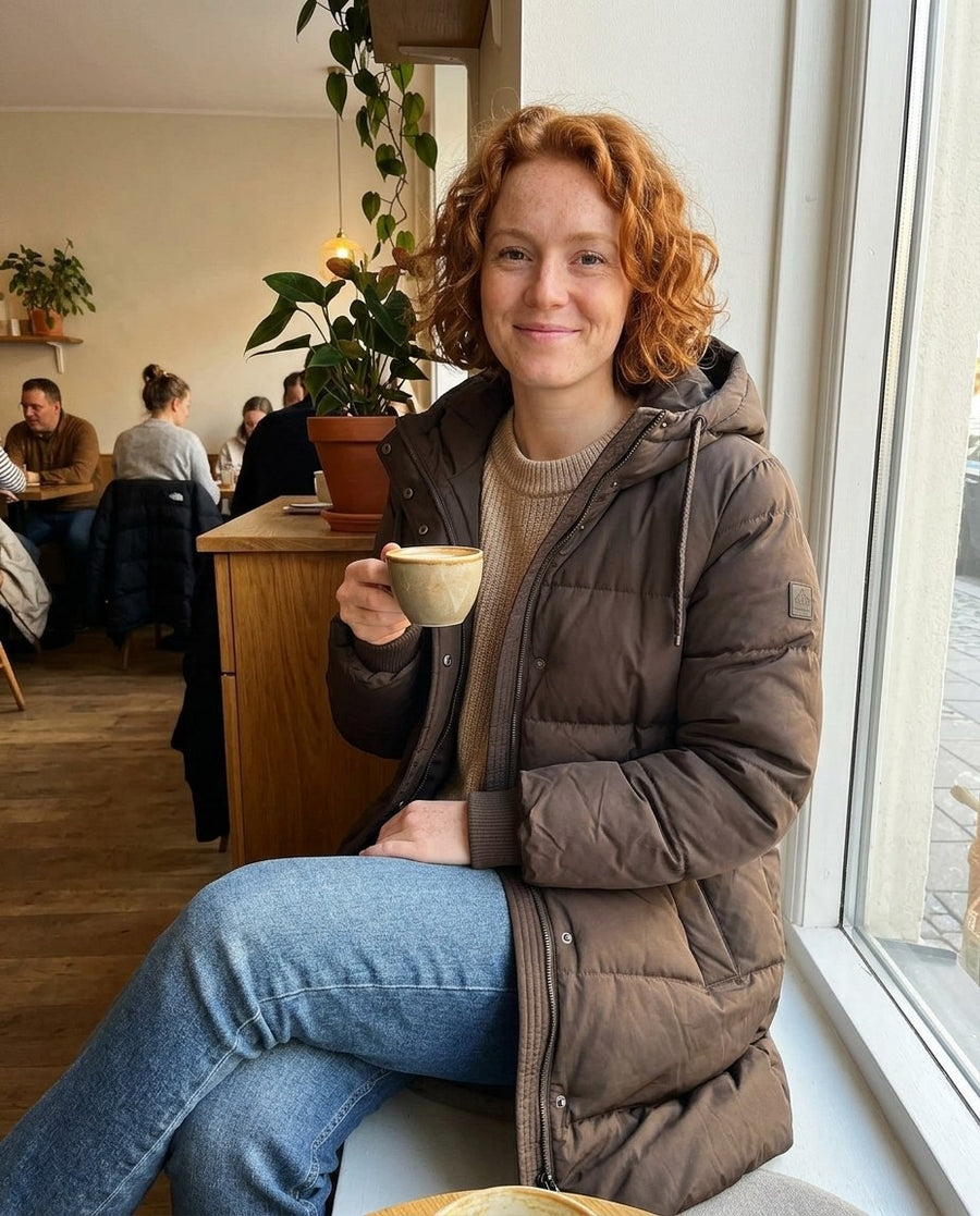 Tyra, lång vadderad jacka med huva för dam i brun, stylinginspiration på ett kafé